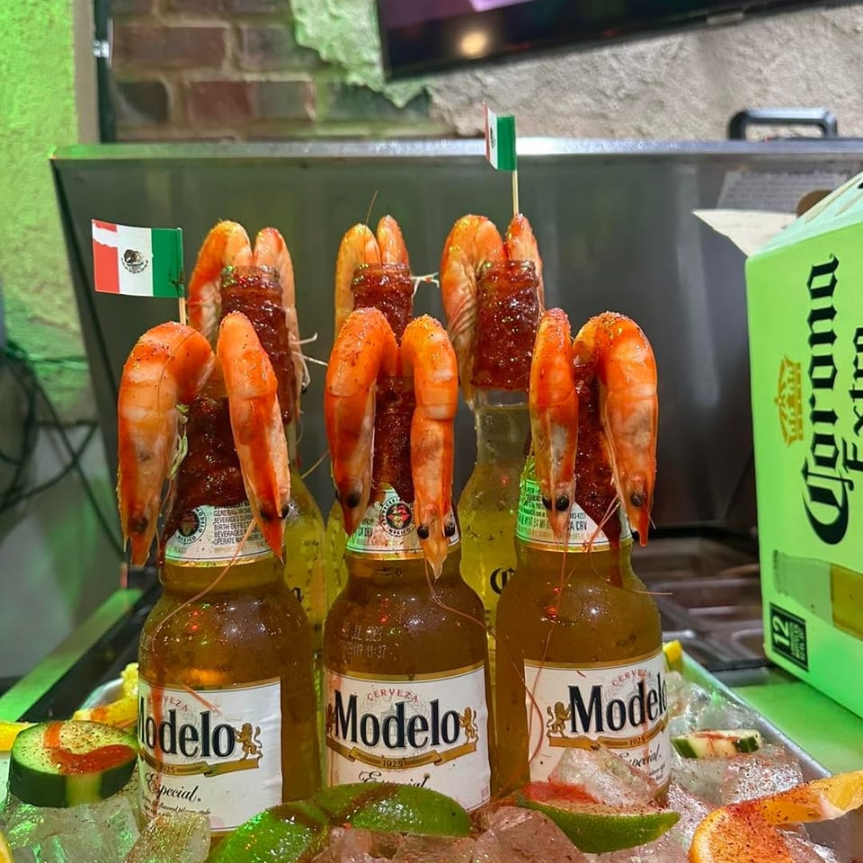 Three Modelo beer bottles topped with grilled shrimp, displayed over ice with lime wedges and spicy garnishes.