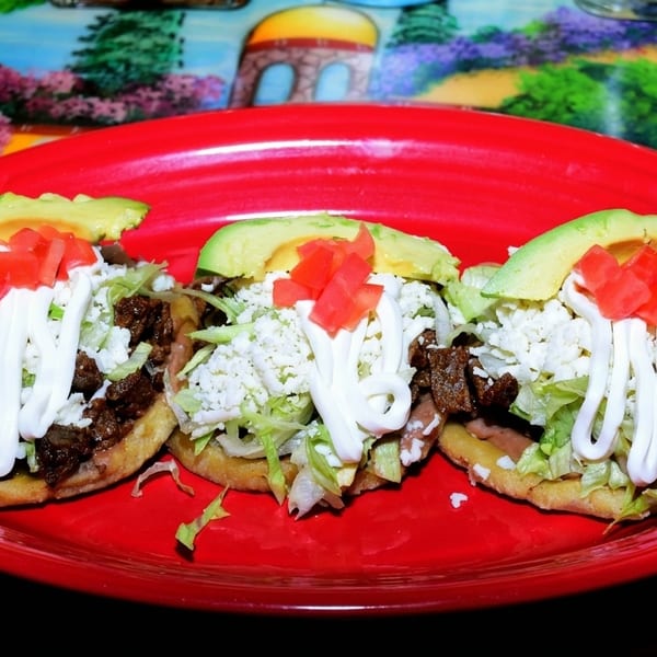 Three sopes with meat, lettuce, cheese, sour cream, diced tomatoes, and avocado on a red plate.