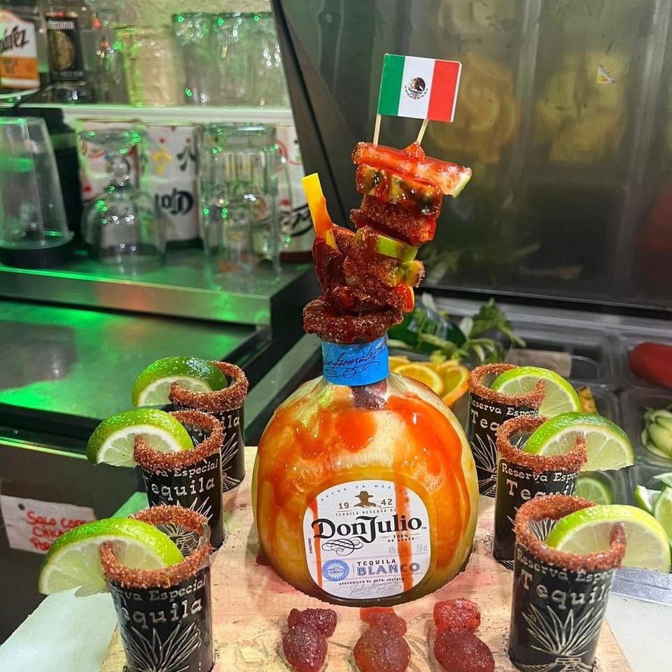 Don Julio tequila bottle surrounded by chili-rimmed tequila cups, lime wedges, garnishes, and a Mexican flag-topped skewer.