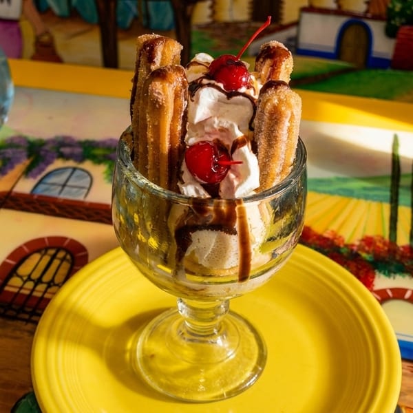 Churro sundae with whipped cream, chocolate syrup, and cherries on a yellow plate with a tequila bottle.