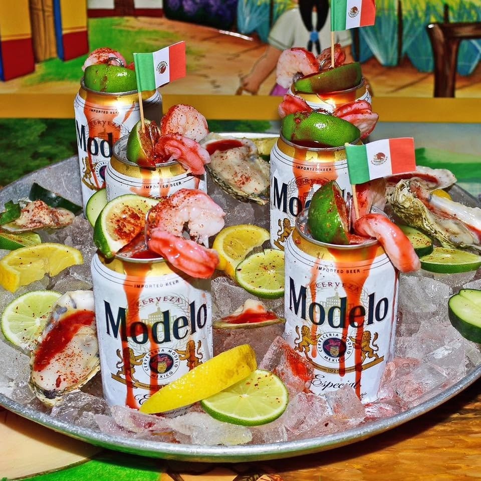 Chilled Modelo beers topped with shrimp, lime, cucumber, and Mexican flags on an ice-filled seafood platter.