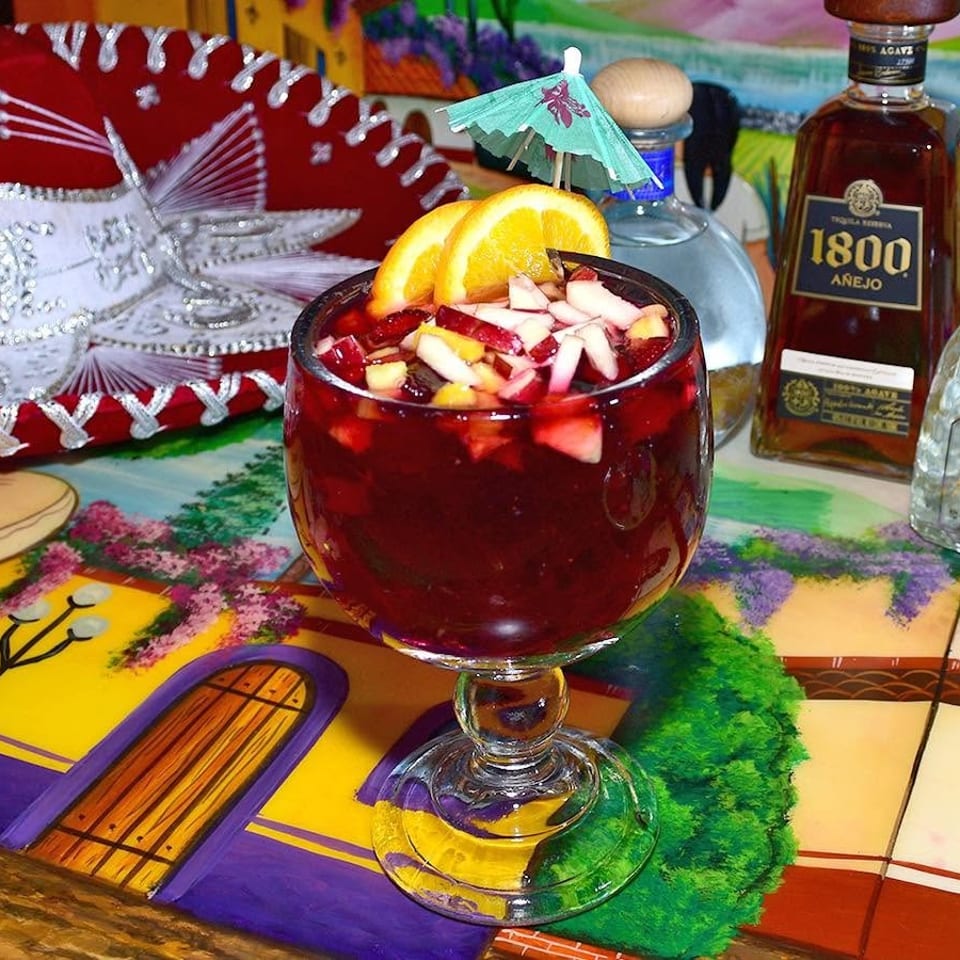 Red fruit cocktail garnished with orange slices sits before tequila bottles, a sombrero, and colorful Mexican décor.