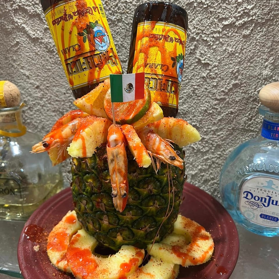 Mexican seafood platter with shrimp, pineapple, citrus, hot sauce, and two bottled beverages surrounding a Mexican flag pick.