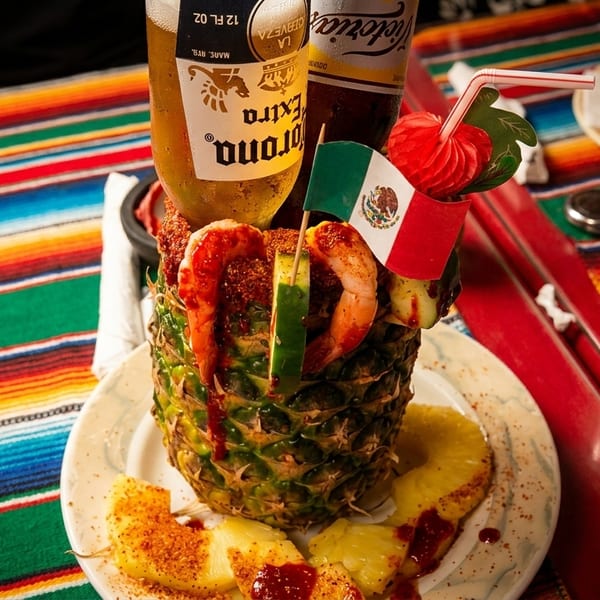 A pineapple filled with shrimp, cucumber, and two beer bottles, on a colorful striped tablecloth.