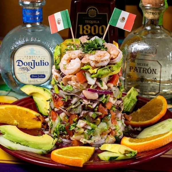 Shrimp ceviche tower with avocado, oranges, and Mexican flags, surrounded by tequila bottles.